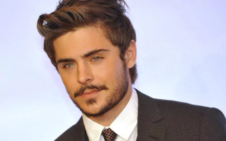 HD PC desktop wallpaper featuring American actor and celebrity Zac Efron with styled hair and a trimmed beard, dressed in a dark suit and tie against a light background.