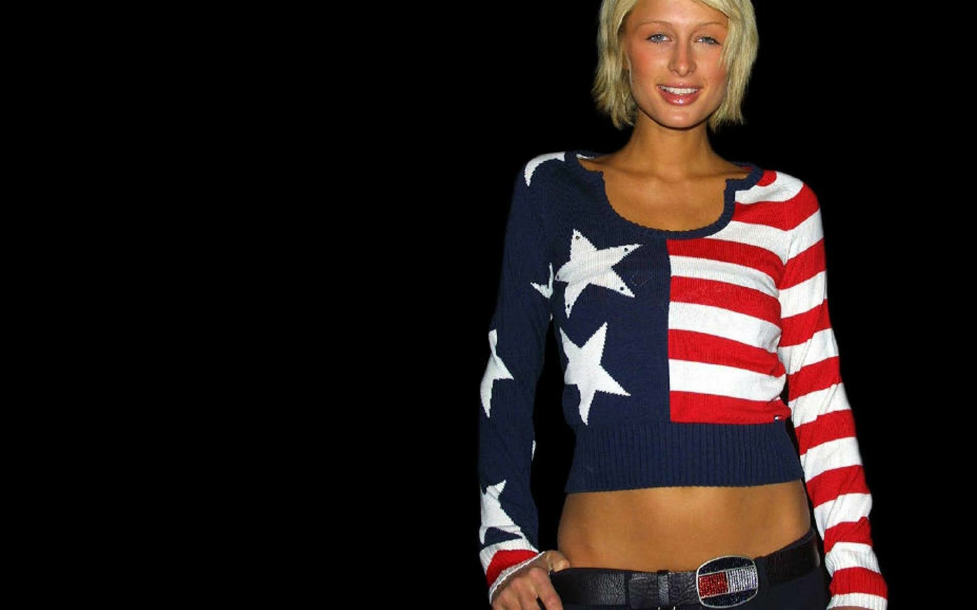 HD PC desktop wallpaper of a celebrity wearing an American-flag crop top and low-rise belt, smiling against a solid black background.