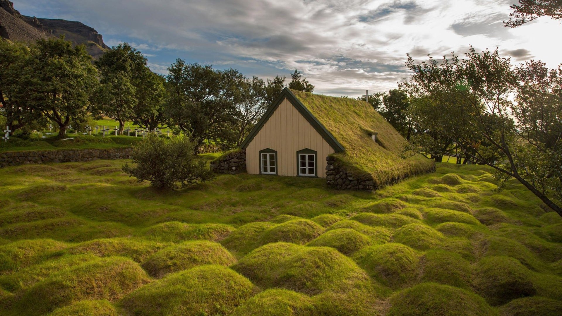 HD desktop wallpaper featuring a quaint church with a grass-covered roof, surrounded by lush green moss and trees under a dramatic cloudy sky.