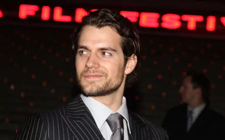 A stylish HD wallpaper featuring British actor Henry Cavill at a film festival, exuding charm and elegance in a sharp suit amidst a vibrant backdrop.