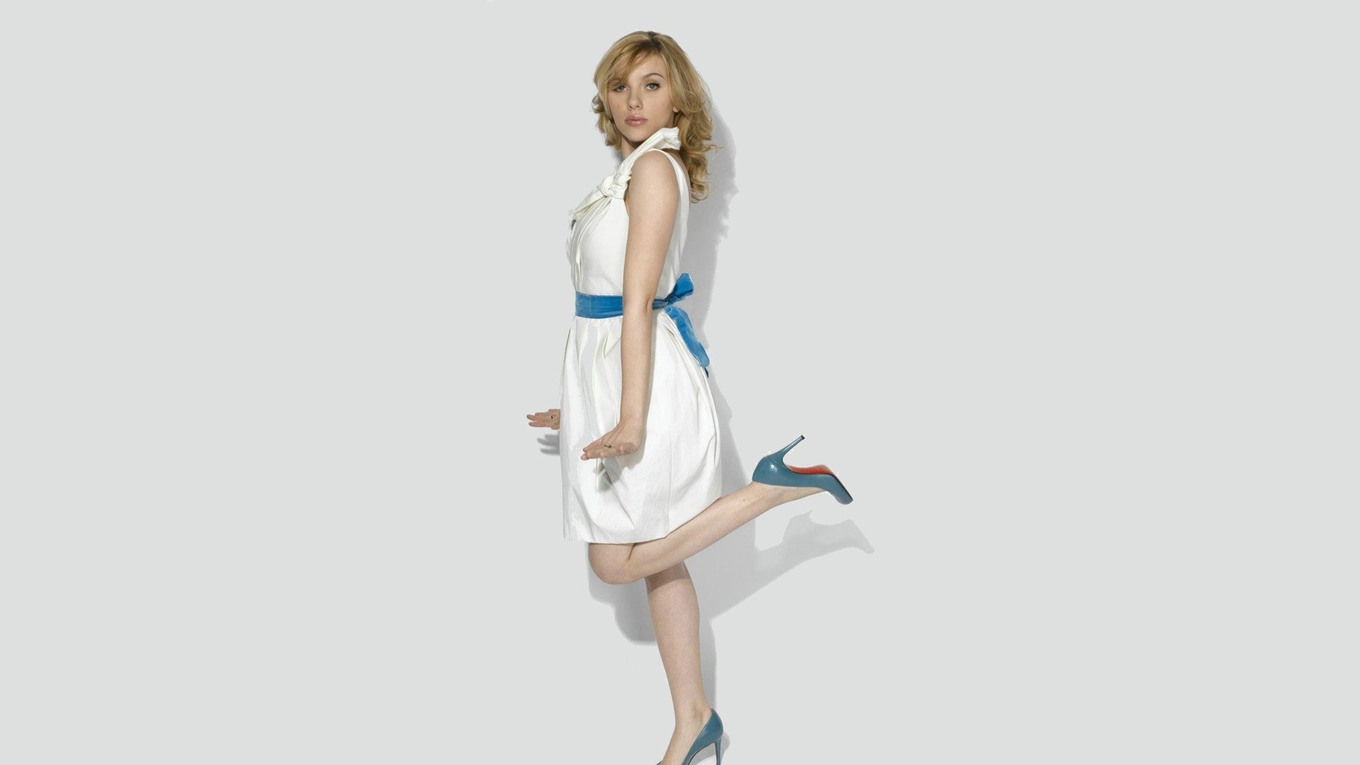 A striking HD desktop wallpaper featuring Scarlett Johansson in a stylish white dress with a blue ribbon, playfully posing with one leg raised.