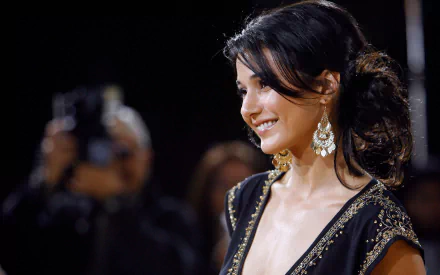 Smiling woman in an elegant black gown with gold embroidery and chandelier earrings, hair in a loose updo, photographed at an evening event — 2K Quad HD desktop wallpaper.