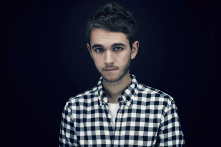 Studio portrait of a young man in a checked shirt, moody lighting suggesting electronic music energy against a dark backdrop — 5K Ultra HD PC desktop wallpaper.