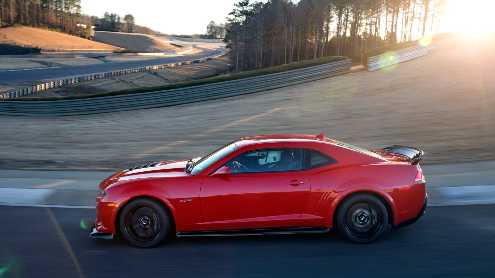 HD PC desktop wallpaper featuring a red Chevrolet Camaro Z28 driving on a sunlit race track surrounded by trees.