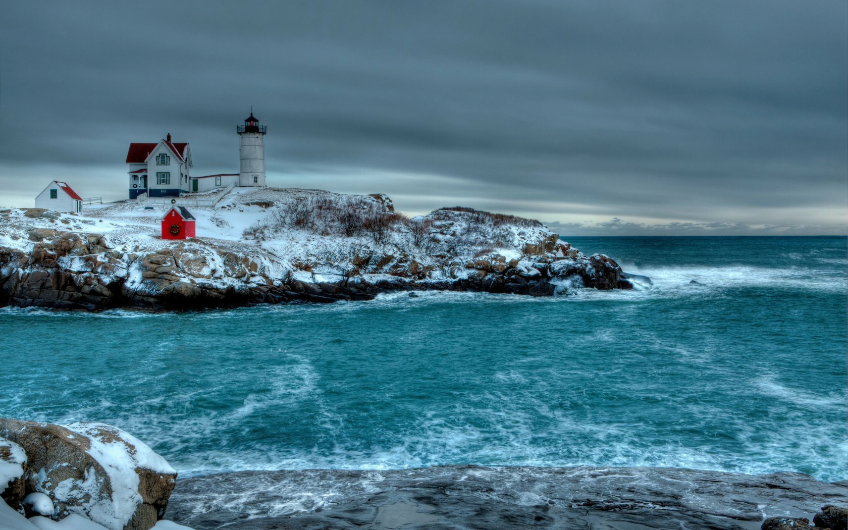 HD desktop wallpaper of a man-made lighthouse and buildings perched on rocky coastline under a cloudy sky with ocean waves crashing.