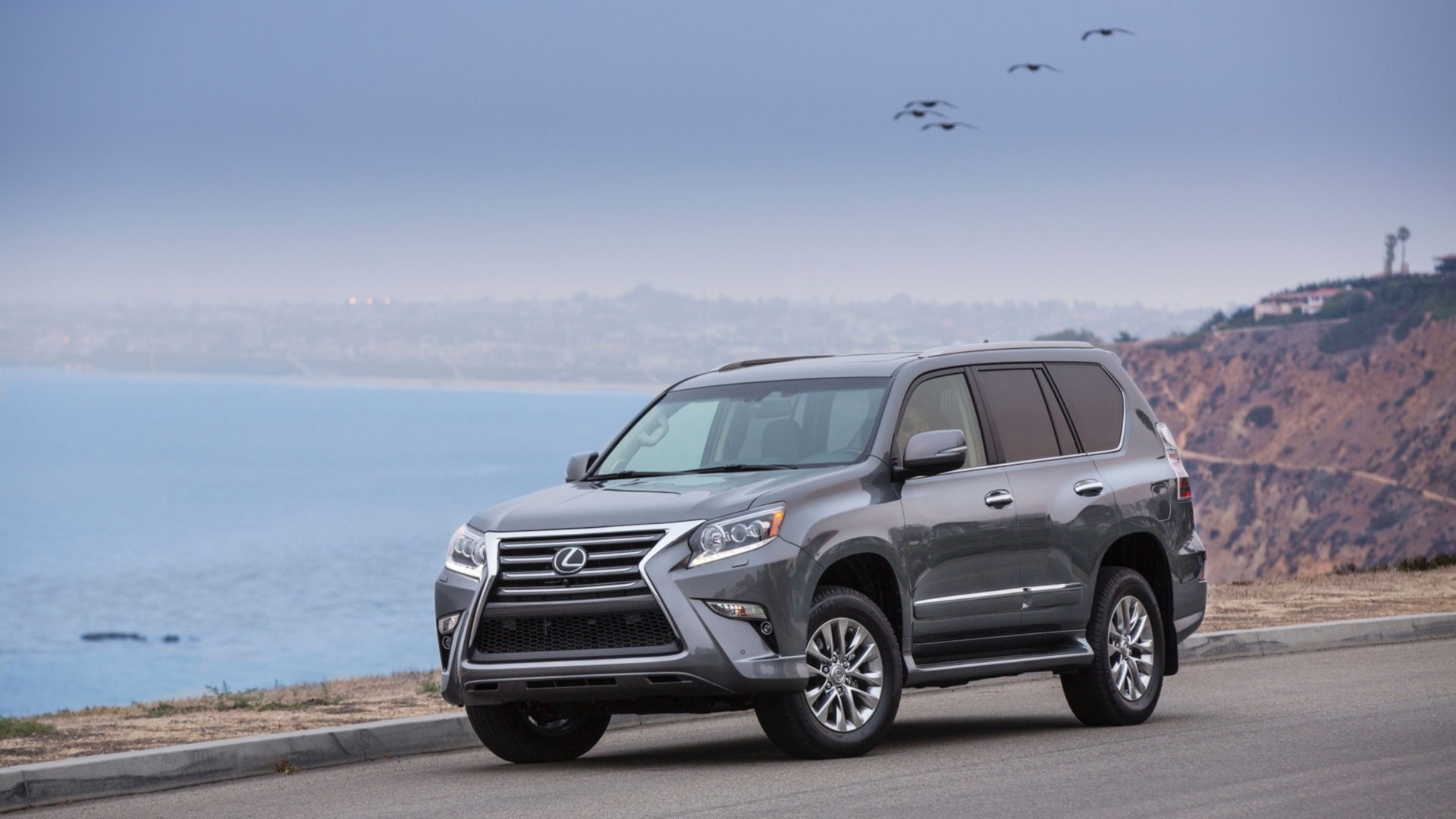 HD PC desktop wallpaper featuring a dark gray Lexus GX parked on a coastal road with cliffs and ocean in the background under a cloudy sky.
