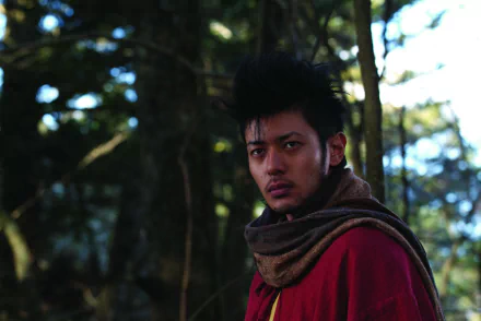 HD PC desktop wallpaper of a brooding shinobi from the movie Shinobi: Heart Under Blade, clad in a red cloak and scarf, standing in a dim forest with an intense, focused stare.