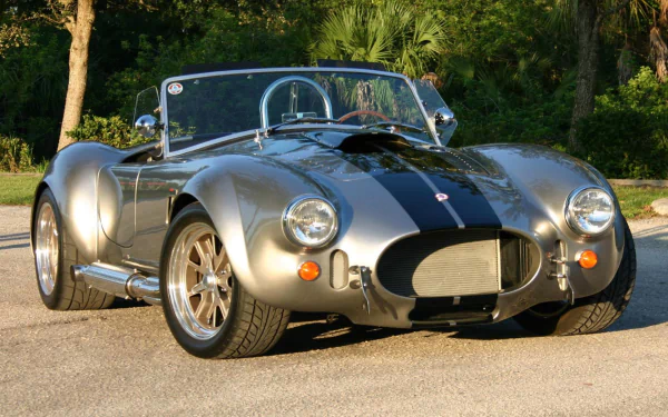 HD desktop wallpaper featuring a classic AC Cobra vehicle parked on a sunlit road surrounded by greenery.