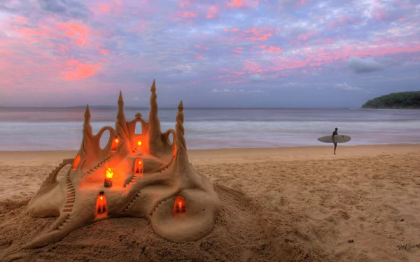 A detailed sandcastle illuminated by small lanterns sits on a sandy beach at sunset, with a surfer walking along the ocean shore under a colorful sky.