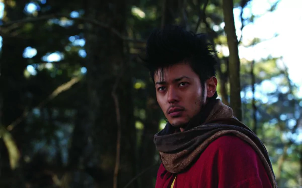 HD PC desktop wallpaper of a brooding shinobi from the movie Shinobi: Heart Under Blade, clad in a red cloak and scarf, standing in a dim forest with an intense, focused stare.
