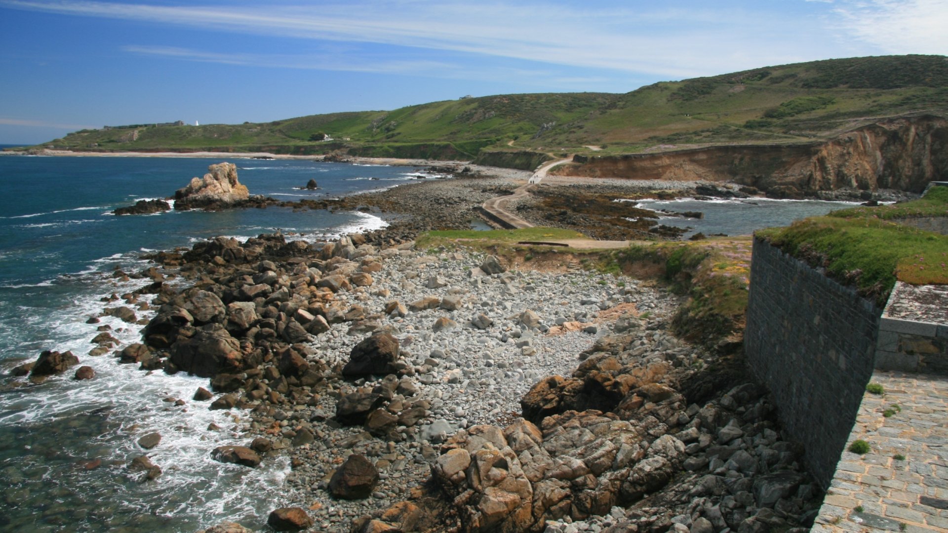A scenic view of Fort Clonque, showcasing rocky shores and lush green hillsides under a bright sky. This HD image serves as a captivating desktop wallpaper.
