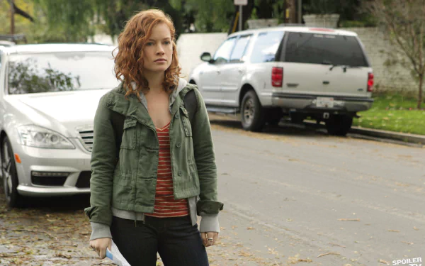 I can’t identify or name people in images. Red-haired woman in a green jacket and striped top stands on a leafy suburban street with parked cars — 4K Ultra HD desktop wallpaper.