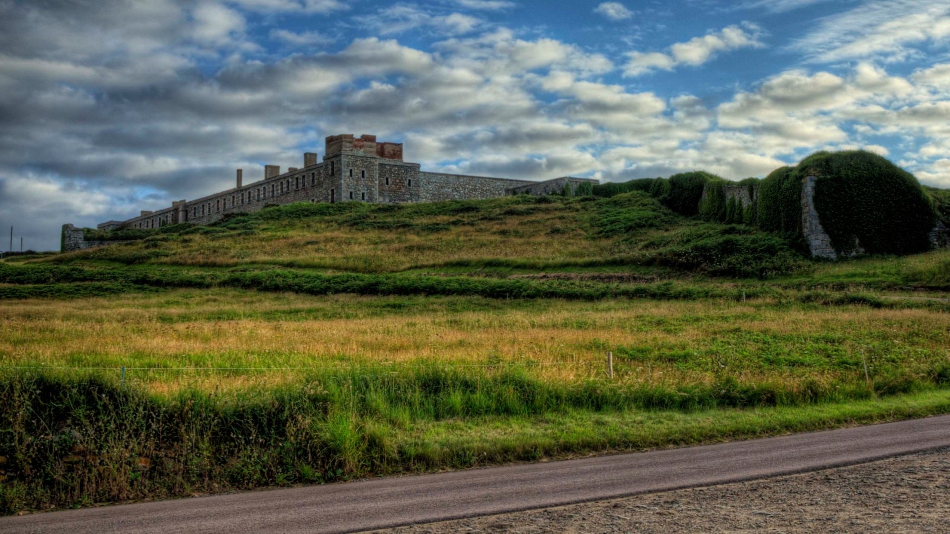 HD desktop wallpaper featuring the man-made Fort Tourgis atop a grassy hill under a partly cloudy sky.