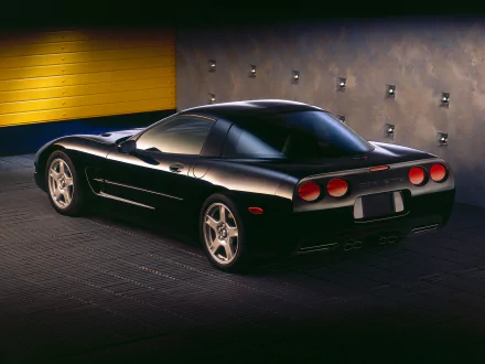 HD desktop wallpaper featuring a sleek black Chevrolet Corvette (C5) parked in an industrial-style garage with yellow and gray walls.