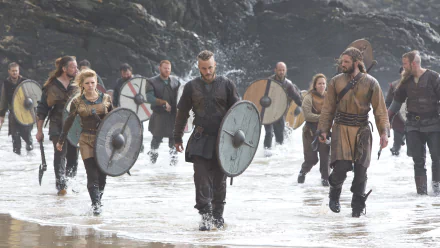 Lagertha and fellow Vikings warriors from the TV show Vikings (2013) march through shallow water, shielded and ready for battle, in a 4K Ultra HD desktop wallpaper background.