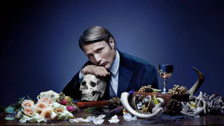 HD PC desktop wallpaper featuring a dark, moody scene from the TV show Hannibal, with a man leaning on a skull surrounded by flowers, horns, and a goblet.