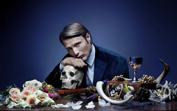 HD PC desktop wallpaper featuring a dark, moody scene from the TV show Hannibal, with a man leaning on a skull surrounded by flowers, horns, and a goblet.