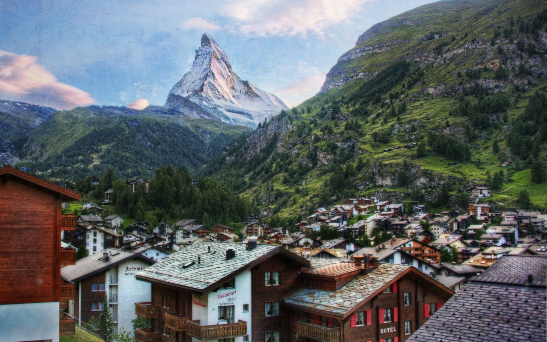 HD PC desktop wallpaper: panoramic view of the Matterhorn rising above a Swiss alpine village, green slopes and traditional chalets under a blue sky.