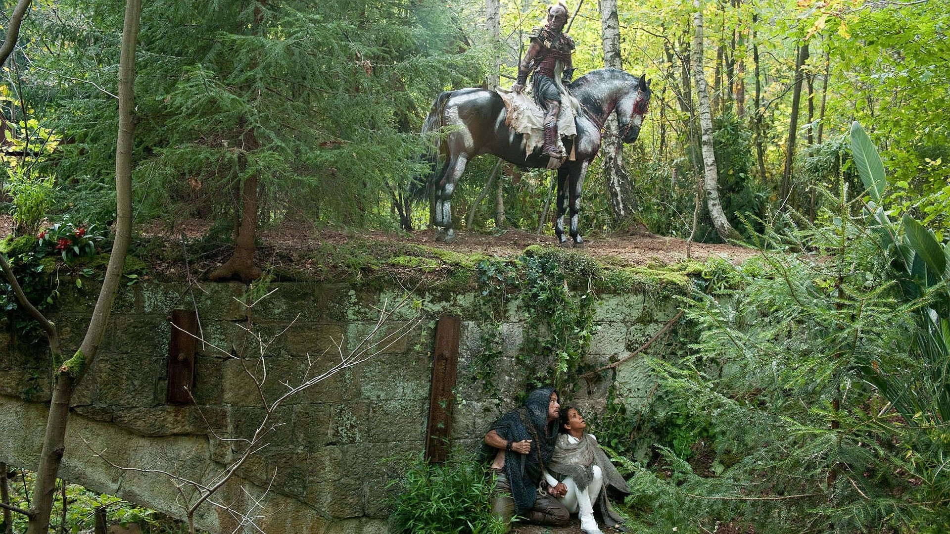 HD PC desktop wallpaper from the movie Cloud Atlas: armored rider on horseback watches from a mossy cliff as two figures shelter below in a lush green forest.