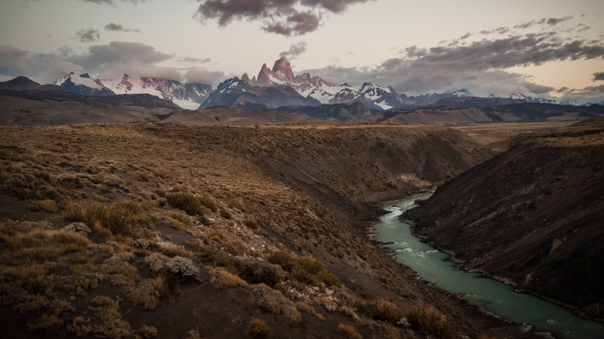 HD PC desktop wallpaper: sweeping Patagonian plain with a turquoise river carving a canyon toward jagged Mount Fitz Roy beneath moody clouds.