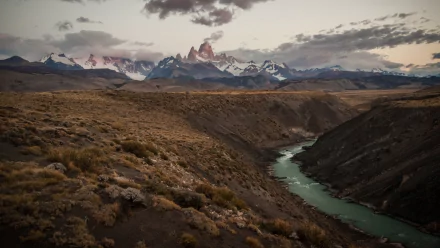 HD PC desktop wallpaper: sweeping Patagonian plain with a turquoise river carving a canyon toward jagged Mount Fitz Roy beneath moody clouds.