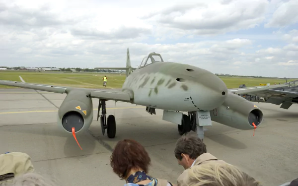 HD desktop wallpaper featuring a Messerschmitt Me 262 military aircraft displayed outdoors on a runway under a cloudy sky.