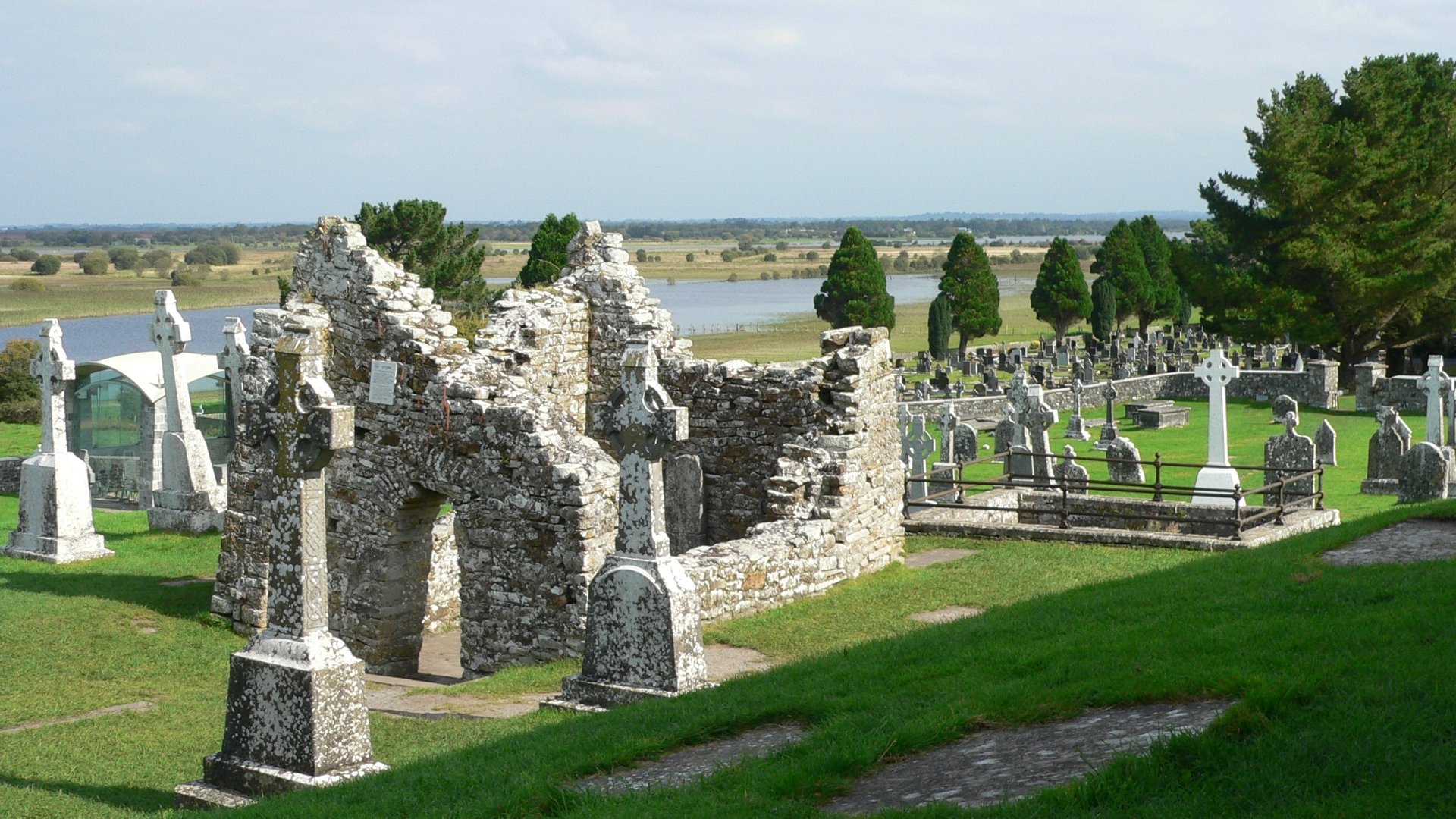 Clonmacnoise Monastery ruins and graveyard with stone crosses on a green Irish riverside landscape — religious medieval architecture captured as a 2K Quad HD PC desktop wallpaper.