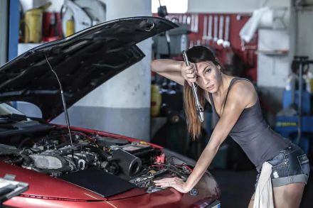 A confident woman leans over a car engine, holding a wrench, set in a garage. This HD desktop wallpaper captures the dynamic theme of women and cars.