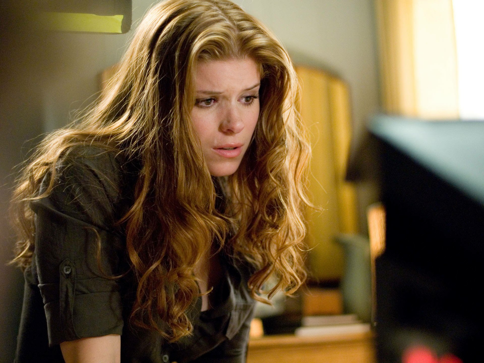 HD PC desktop wallpaper of a celebrity actress with long wavy auburn hair leaning toward a monitor, wearing a dark jacket and gazing intently at the screen.