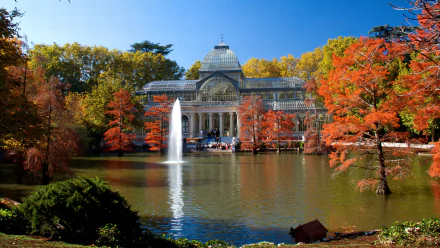 HD desktop wallpaper featuring the Palacio de Cristal surrounded by autumn trees and a pond with a fountain in a serene, man-made park setting.