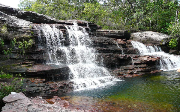 Caño Cristales waterfall cascading over layered rocks into a clear pool surrounded by lush forest — vibrant nature scene, 2K Quad HD PC desktop wallpaper and background.