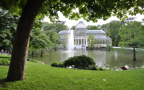 HD desktop wallpaper of the man-made Palacio de Cristal, framed by lush green trees and a pond with a central water fountain in a serene park setting.