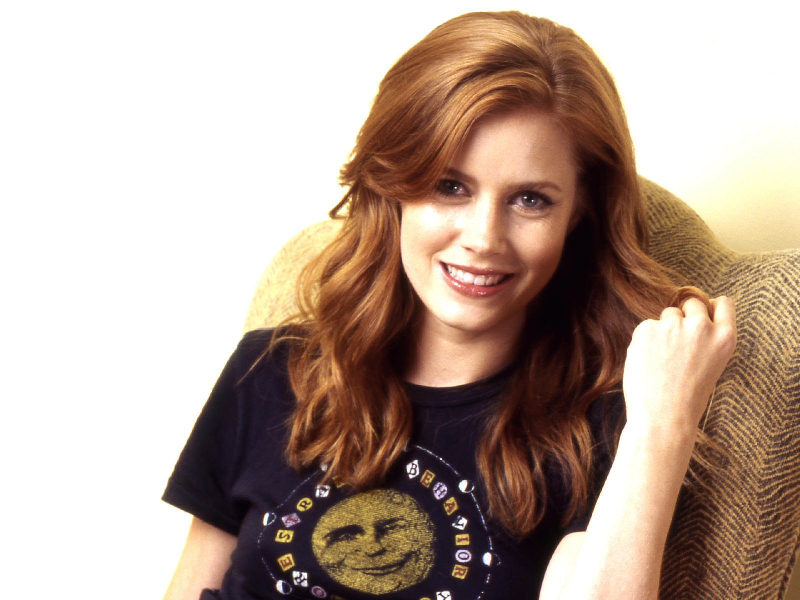 HD desktop wallpaper featuring a smiling woman with red hair, seated in a beige chair, wearing a black t-shirt.