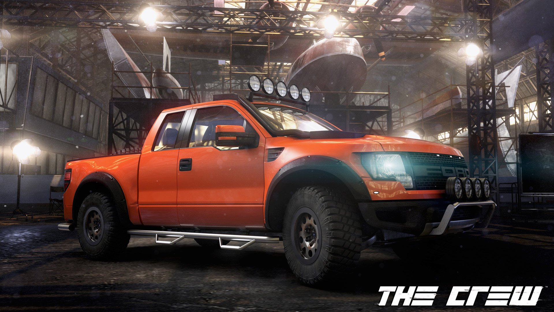 HD PC desktop wallpaper featuring a bright orange off-road truck from the video game The Crew, set in an industrial warehouse environment.