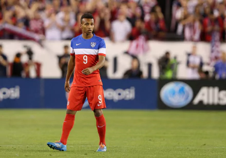 HD wallpaper of Julian Green in a blue and red soccer kit on the field, poised for action, with a blurred stadium background.