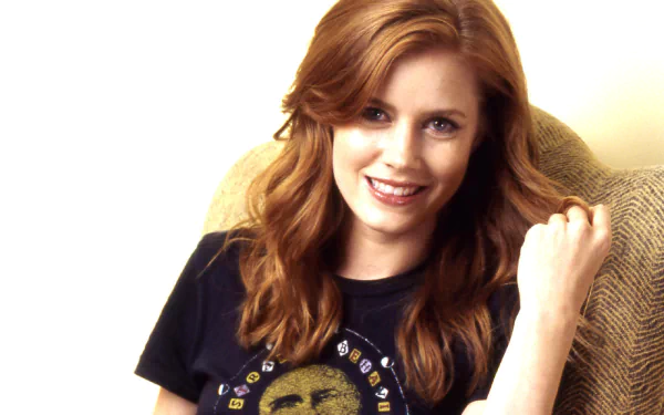 HD desktop wallpaper featuring a smiling woman with red hair, seated in a beige chair, wearing a black t-shirt.
