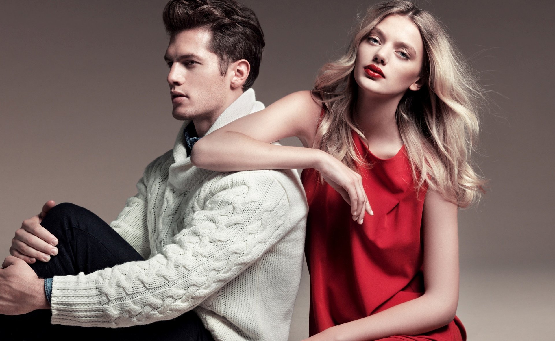 HD desktop wallpaper of two fashion models: a Dutch blonde woman in a red dress leaning on a seated man in a white cable‑knit sweater against a neutral studio background.