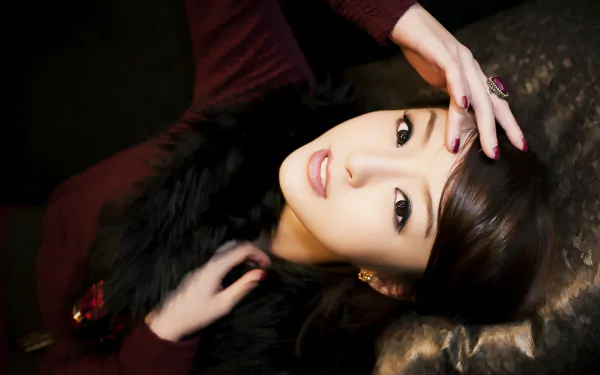 HD wallpaper of an Asian woman with dark hair and expressive eyes. She is relaxing, looking upwards with one hand on her forehead, wearing a burgundy top and a dark fur stole.