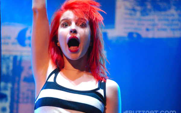Red‑haired rock singer in striped top, mouth open mid-song onstage; vibrant, energetic 2K Quad HD PC desktop wallpaper evoking Paramore's alt‑rock music.