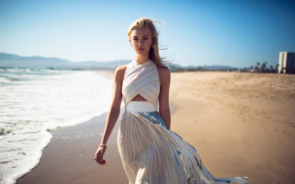 A stunning HD desktop wallpaper featuring Nicola Peltz walking along a beach in a flowing dress, against a backdrop of waves and blue skies.