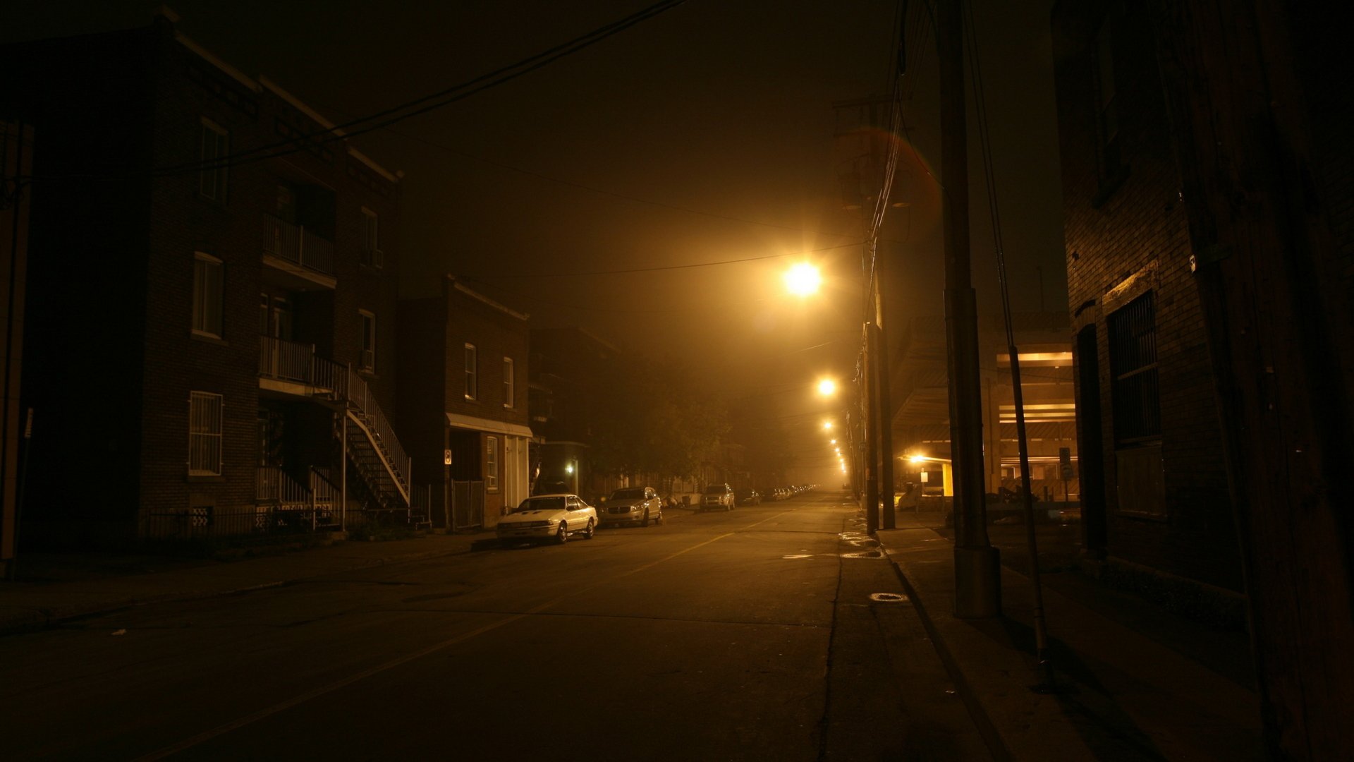 HD desktop wallpaper of a dimly lit town street at night with glowing streetlights and parked cars, showcasing a quiet, man-made urban scene.