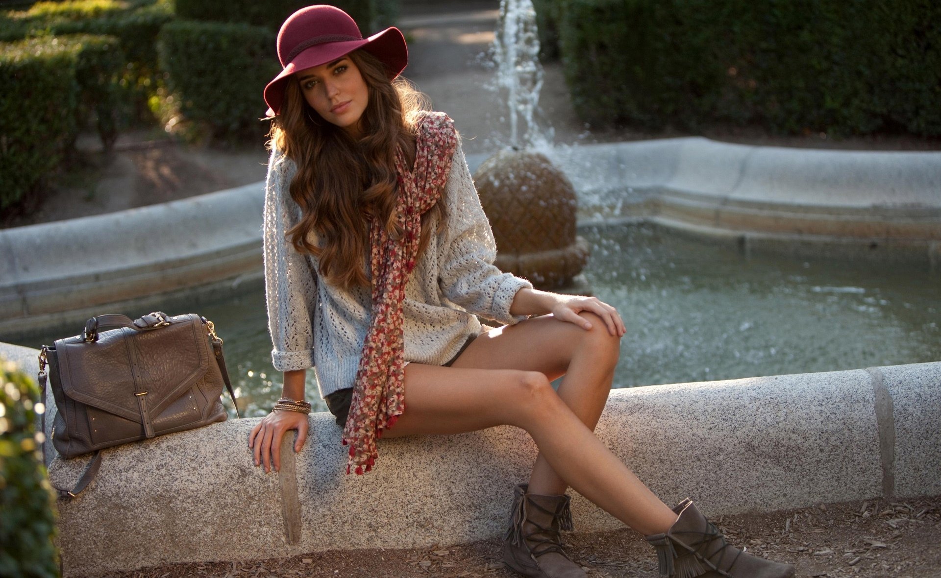 A stylish Spanish woman, Clara Alonso, poses elegantly by a tranquil fountain, wearing a hat and patterned scarf, set against a lush garden backdrop. HD desktop wallpaper.