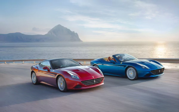 Two Ferrari California T vehicles, one red and one blue, drive along a coastal road with mountains and ocean in the background, captured in an HD PC desktop wallpaper.
