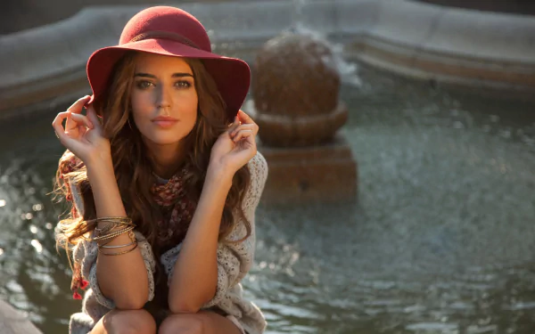 A Spanish woman, Clara Alonso, poses gracefully by a fountain, wearing a maroon hat and a cozy sweater, captured in a beautiful HD desktop wallpaper.