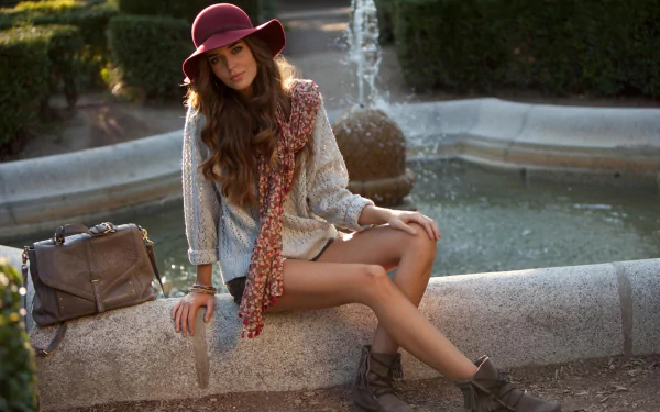 A stylish Spanish woman, Clara Alonso, poses elegantly by a tranquil fountain, wearing a hat and patterned scarf, set against a lush garden backdrop. HD desktop wallpaper.