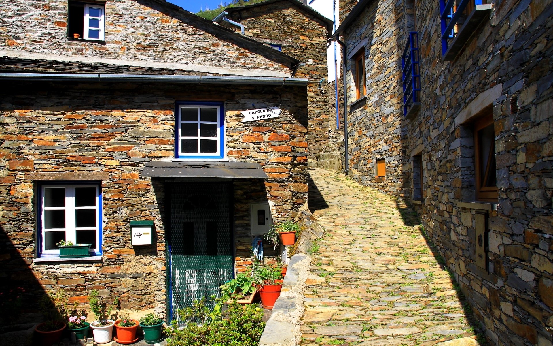 HD PC desktop wallpaper/background showing sunlit stone houses and a narrow cobblestone alley with potted plants — man-made rustic house facades and blue-trimmed windows.