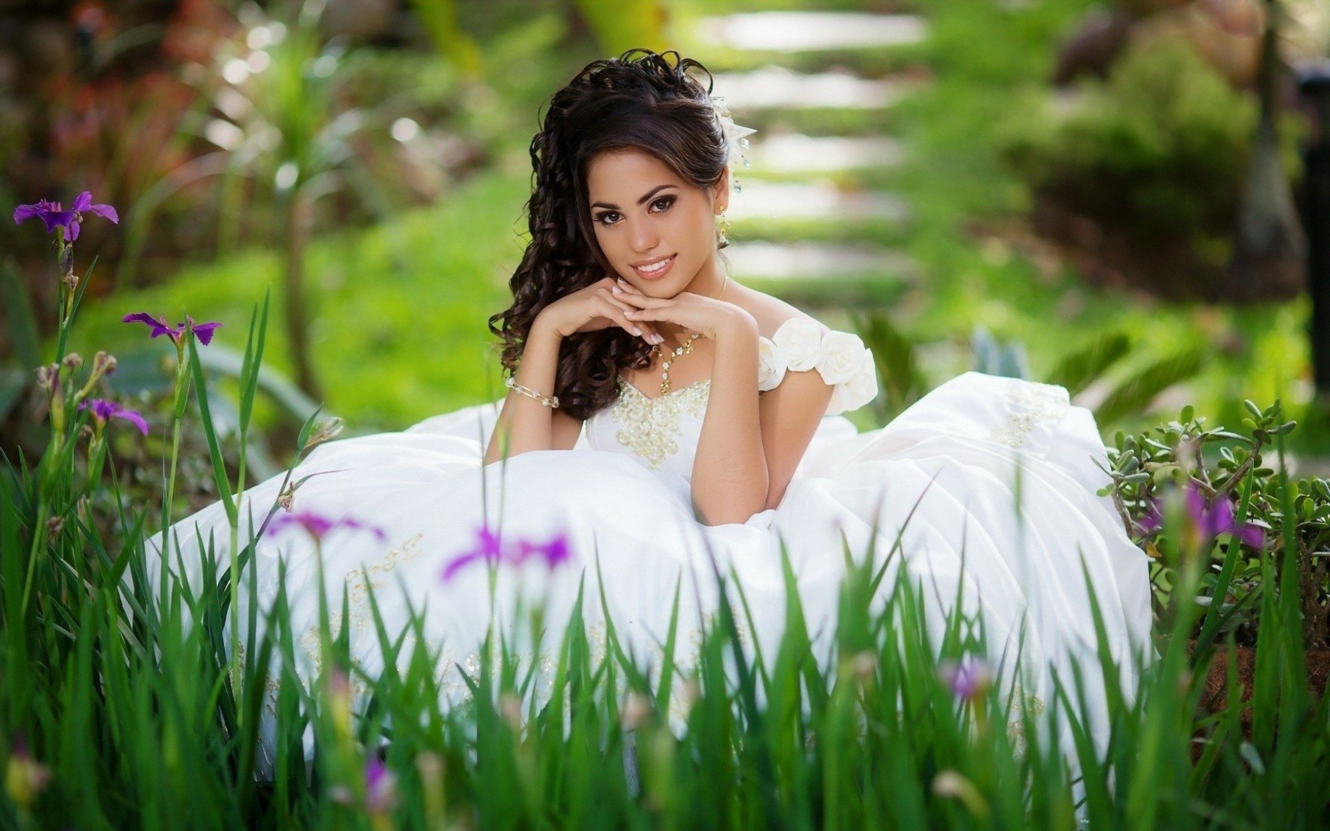 A bride in a white dress smiles while sitting in a lush garden, captured in a vibrant HD PC desktop wallpaper and background.