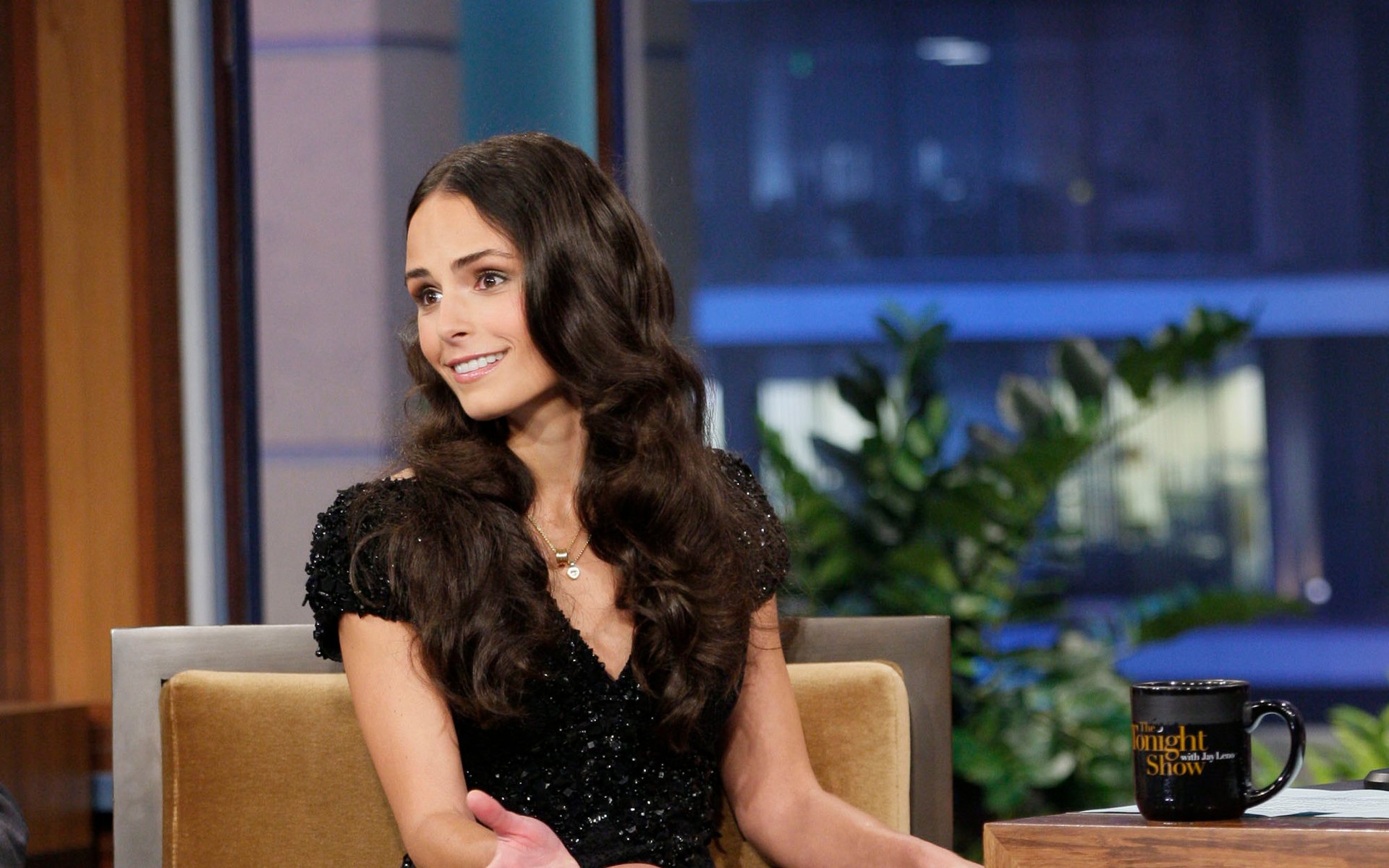 Smiling woman with long dark hair in a black dress seated on a talk-show couch, studio backdrop with plants and a coffee mug; HD desktop wallpaper.