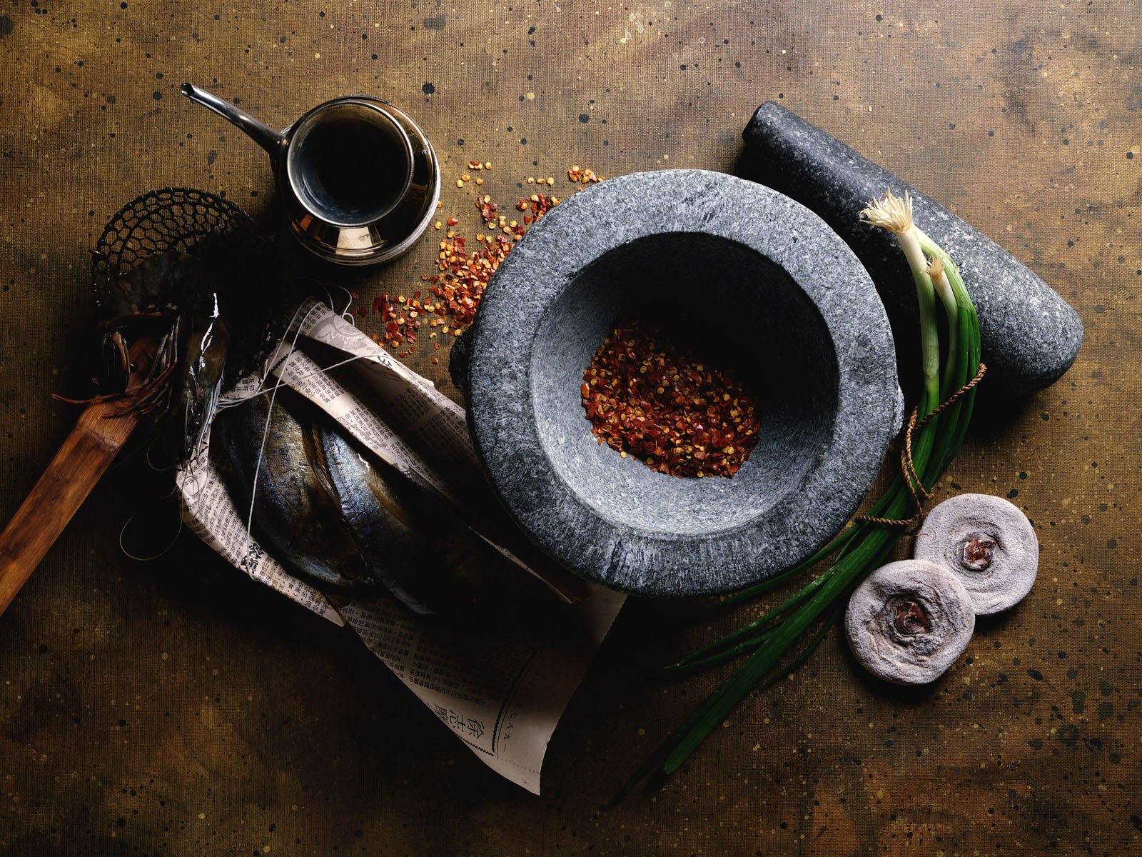 HD PC desktop wallpaper showing spices and food: a stone mortar with crushed red pepper flakes, garlic cloves, green onions and kitchen tools on a rustic surface.