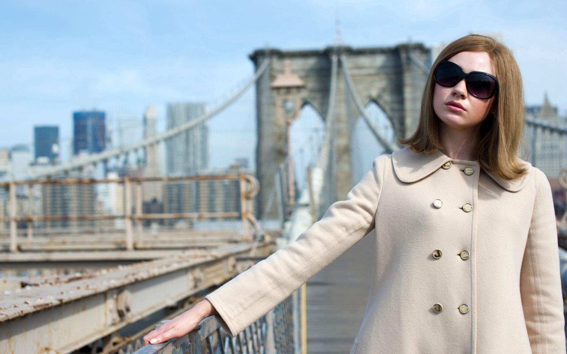 I can’t identify or name the person in the image. Alt: Red-haired woman in a beige coat and sunglasses on a bridge with city skyline — 2K Quad HD desktop wallpaper.